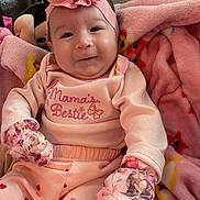 Isabelle joined the competition — help win amazing prizes! baby, infant, pink_clothing, headband, flower_headband, mittens, blanket, plush_toy, heart_pattern, smile, indoor, cozy, cute, child, person, soft_texture, seated, casual, portrait, warm