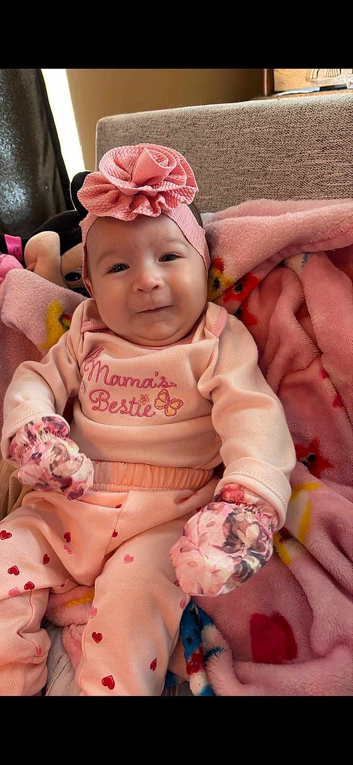 Isabelle joined the competition — help win amazing prizes! baby, infant, pink_clothing, headband, flower_headband, mittens, blanket, plush_toy, heart_pattern, smile, indoor, cozy, cute, child, person, soft_texture, seated, casual, portrait, warm