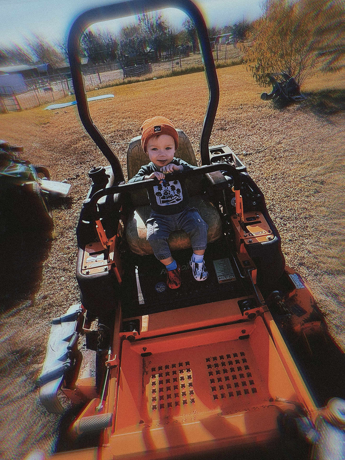 Cooper joined the competition — help win amazing prizes! child, toddler, orange_beanie, lawn_mower, outdoor, grass, fence, sunny, equipment, trees, seat, steering_bar, footwear, playful, yard, person, smile, clothing, daylight, rustic