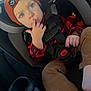 toddler, child, car_seat, beanie, orange_hat, blue_eyes, finger_in_mouth, red_plaid_shirt, brown_pants, car_interior, seatbelt, safety_harness, curious_expression, casual_clothing, cute, indoors, young_child, portrait, infant, baby