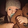 baby, child, pacifier, hat, knit_hat, sweater, teddy_bear, plush_toy, close_up, cute, infant, hand, face, portrait, warm, soft_texture, cozy, indoor, expression, young