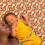 Reign is registered to the contest to win money with this photo: child, adult, smiling, toddler, yellow_outfit, patterned_blanket, orange, white, playing, happy, family, lying_down, closeup, skin, person, casual_clothing, relaxing, indoor, affection, bonding