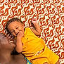 child, adult, smiling, toddler, yellow_outfit, patterned_blanket, orange, white, playing, happy, family, lying_down, closeup, skin, person, casual_clothing, relaxing, indoor, affection, bonding