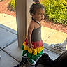 toddler, child, dress, colorful_clothing, green_sneakers, outdoor, sidewalk, pillar, plants, sunny, curly_hair, casual, person, young_child, daylight, shoes, standing, curious, hair_tie, concrete