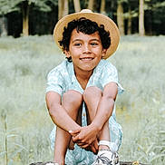 Malakai is registered to the contest to win money with this photo: boy, casual_clothing, child, curly_hair, daylight, grass, greenery, happy, hat, nature, outdoor, patterned_shoes, portrait, shoes, short_sleeves, sitting, smiling, straw_hat, tree_stump, white_shoes