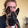 animal, black_shirt, closeup, cute, dog, ear, face, fur, german_shepherd, hand, holding, home, indoor, person, pet, portrait, puppy, tattoo, wall, young_adult