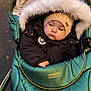 baby, child, pram, stroller, jacket, hood, hat, knit_hat, fur_trim, outdoor, autumn, fall_leaves, green, black_clothing, cute, cold_weather, cozy, portrait, infant, person