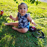 Amelia is registered to the contest to win money with this photo: baby, child, grass, outdoor, sunlight, smile, camera, leaf_pattern, blue_clothing, nature, greenery, sitting, happy, face, person, summer, park, toy, cute, eyes