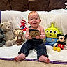 alien_toy, baby, blanket, carpet, child, couch, cute, denim_overalls, happy, holding, indoor, infant, lion_plush, mickey_mouse, plush_toys, sitting, smiling, spiderman_toy, teddy_bear, wooden_plaque