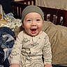 baby, infant, smiling, dinosaur_print, onesie, beanie, bed, blanket, pillow, happy, child, indoors, cute, person, sleepwear, furniture, cozy, portrait, sitting, home