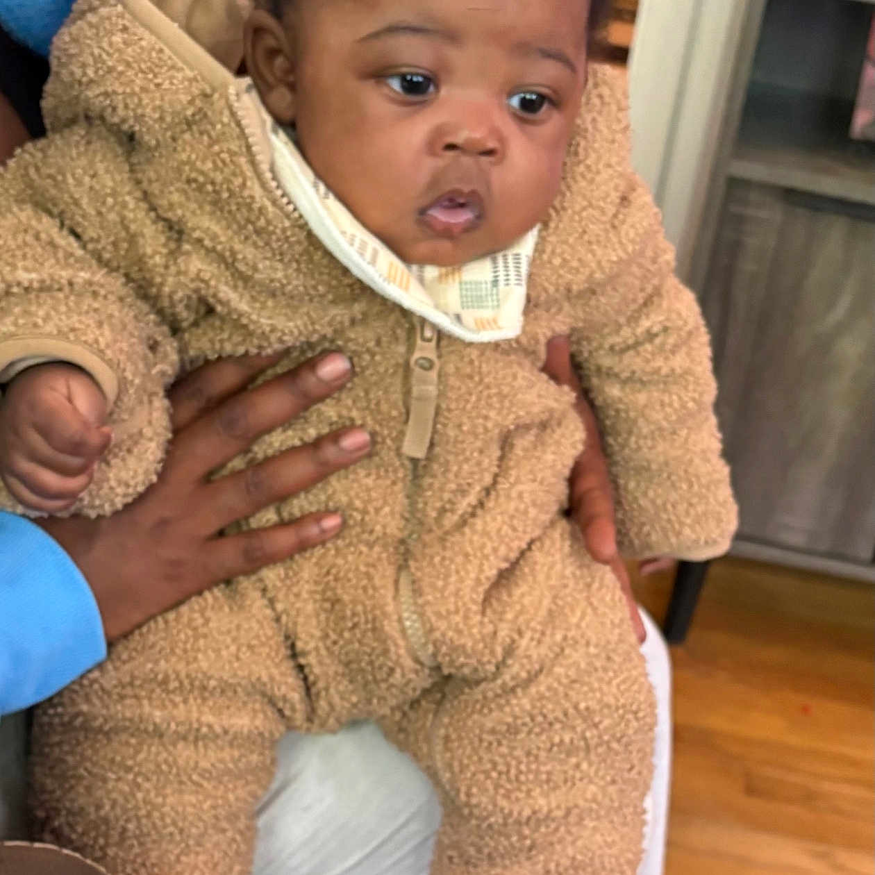 Legend joined the competition — help win amazing prizes! baby, infant, child, fuzzy_clothing, onesie, indoor, person, hand, curly_hair, seated, wooden_floor, furniture, adult_hand, warm_lighting, cute, cozy, expression, face, socks, holding