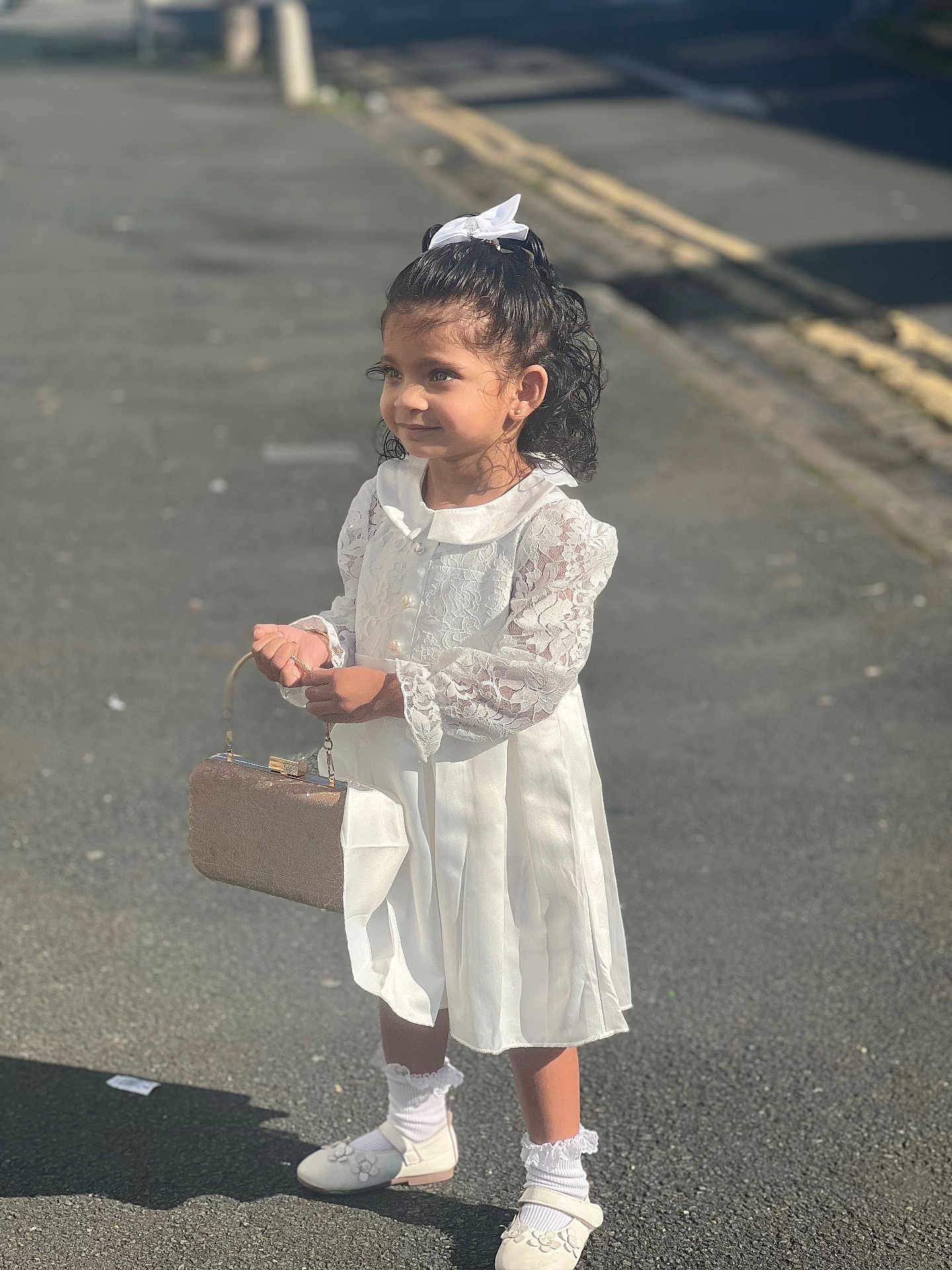 Daleyza is registered to the contest to win money with this photo: child, girl, white_dress, purse, curly_hair, white_bow, street, shoes, socks, sunlight, side_view, smile, outdoor, young_child, fashion, accessories, shadow, pavement, innocence, cute