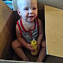 baby, blond_hair, bottle, cardboard_box, casual, child, container, cute, face, hand, happy, indoors, person, red_trim, sitting, skin, smiling, tank_top, toddler, toy