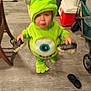 child, toddler, costume, green, monster, indoor, floor, shoes, holding, toy, blurred, person, cute, clothing, footwear, home, baby, standing, carpet, furniture