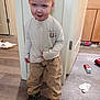 cabinet, child, clothing, door, expression, floor, footwear, hair, indoor, light, messy, pants, paper, person, playful, room, shirt, shoes, standing, toddler