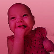 Maja is registered to the contest to win money with this photo: adorable, baby, cheeky, child, closeup, cute, expression, face, hand_in_mouth, happy, head, indoors, infant, pink_light, portrait, skin, sleeveless, smiling, wearing_dress, young_child