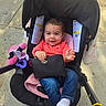 baby, black, blanket, child, clothing, face, footwear, handbag, happy, jeans, outdoor, person, pink, sidewalk, smile, socks, stroller, sunlight, toy, wristlet