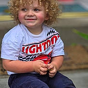 Jaxson is registered to the contest to win money with this photo: child, toddler, curly_hair, smiling, barefoot, sitting, tshirt, pants, hands, face, rosy_cheeks, concrete_floor, toy, indoor, cute, happy, young_child, casual_clothing, lightning_mcqueen, portrait