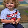 child, toddler, curly_hair, smiling, barefoot, sitting, tshirt, pants, hands, face, rosy_cheeks, concrete_floor, toy, indoor, cute, happy, young_child, casual_clothing, lightning_mcqueen, portrait