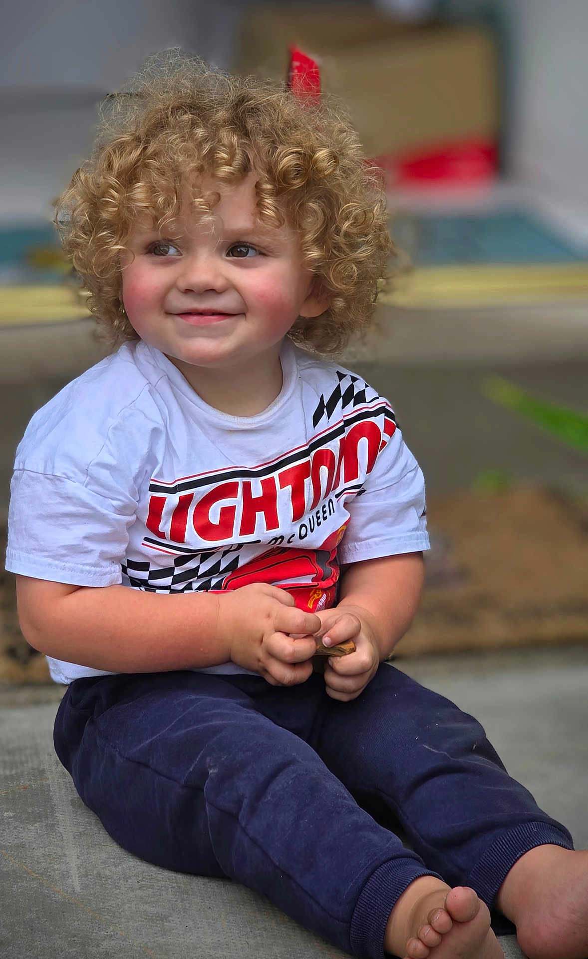 Jaxson is registered to the contest to win money with this photo: child, toddler, curly_hair, smiling, barefoot, sitting, tshirt, pants, hands, face, rosy_cheeks, concrete_floor, toy, indoor, cute, happy, young_child, casual_clothing, lightning_mcqueen, portrait