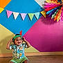 toddler, child, smiling, sitting, floor, headband, feather, colorful, decorations, wall_art, pennant_flags, rainbow_fan, shirt, shorts, indoor, playful, happy, wooden_floor, portrait, casual_clothing