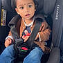 child, toddler, car_seat, seat_belt, brown_jacket, blue_jeans, sneakers, indoors, car_interior, face, curious, bindi, safety, seat, young_child, person, casual_clothing, cute, portrait, baby