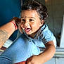 toddler, child, smiling, curly_hair, blue_shirt, hand, adult_arm, tattoo, indoor, floor_tiles, jeans, playful, happy, person, skin, face, tongue_out, holding_on, casual_clothing, close_up