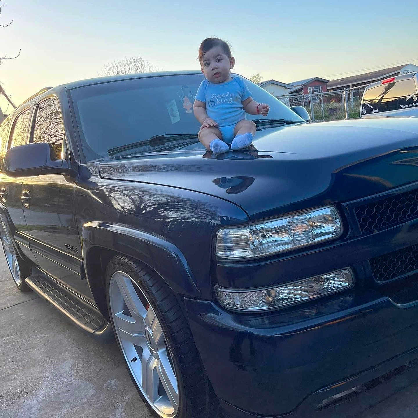 Ximena is registered to the contest to win money with this photo: baby, blue, car, child, curious, evening, hood, metal, onesie, outdoor, person, reflection, residential, sidewalk, sky, sunset, suv, tire, vehicle, wheel
