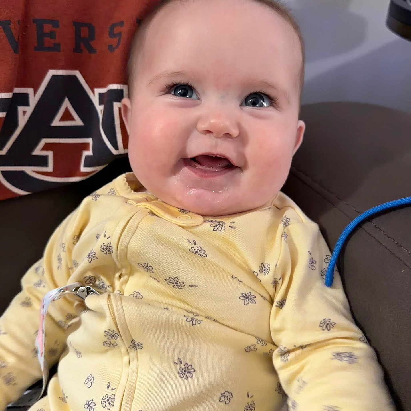 Annie-Ruth is registered to the contest to win money with this photo: baby, blue_eyes, chubby_cheeks, closeup, clothing, couch, cute, drool, fabric, floral_pattern, happy, indoor, infant, pacifier_clip, person, portrait, sitting, smiling, soft_lighting, yellow_onesie