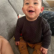 Kaine is registered to the contest to win money with this photo: baby, child, smiling, happy, sitting, indoor, clothing, pants, sweater, dinosaur_pattern, couch, furniture, skin, hand, foot, face, eyes, hair, cozy, comfort