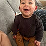 baby, child, smiling, happy, sitting, indoor, clothing, pants, sweater, dinosaur_pattern, couch, furniture, skin, hand, foot, face, eyes, hair, cozy, comfort