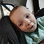 child, car_seat, car_interior, green_shirt, young_child, face, portrait, playful_expression, window, seatbelt, building, daylight, casual_clothing, indoor, person, close_up, head, eyelashes, skin, hair