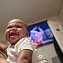 baby, child, smiling, white_shirt, indoor, television, cartoon, happy, holding, person, skin, face, arm, diaper, room, blurred_background, cute, infant, portrait, expression