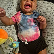 Ja'Leir is registered to the contest to win money with this photo: baby, infant, smiling, tie_dye_shirt, black_shorts, plush_toy, elephant_toy, pillow, patterned_pillow, text_pattern, happy, child, indoors, cute, lying_down, person, mouth_open, joyful, colorful_clothing, soft_toy