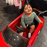 Osvaldo is registered to the contest to win money with this photo: toddler, child, toy_car, red_car, smile, happy, indoor, clothing, floor, steering_wheel, seat, sock, person, shorts, long_sleeve_shirt, play, cute, baby, furniture, expression