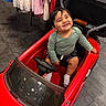 toddler, child, toy_car, red_car, smile, happy, indoor, clothing, floor, steering_wheel, seat, sock, person, shorts, long_sleeve_shirt, play, cute, baby, furniture, expression