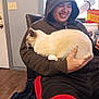 black_pants, bucket, casual, cat, chair, door, furniture, gasoline_can, holding, home, hoodie, indoor, person, pet, red_shorts, relaxed, siamese_cat, smiling, window, wooden_floor