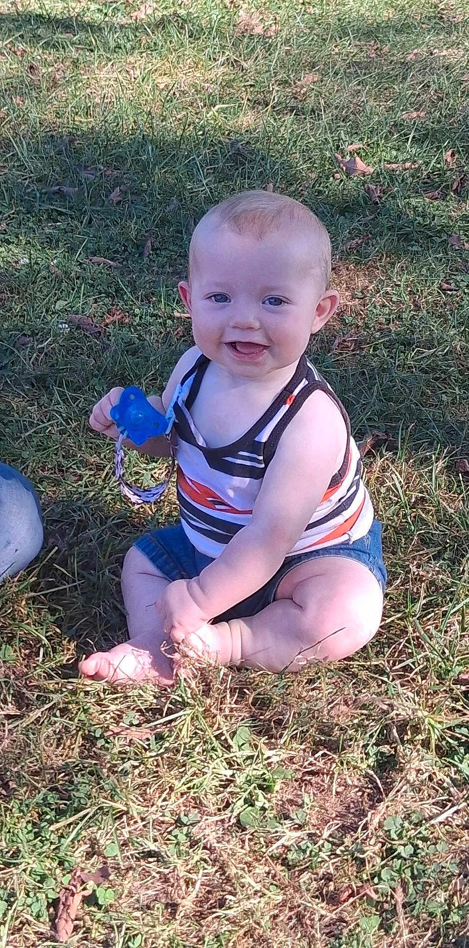 Kolter is registered to the contest to win money with this photo: baby, child, grass, outdoor, smiling, sitting, pacifier, tank_top, denim_shorts, sunlight, happy, cute, person, nature, summer, barefoot, playful, young, infant, casual