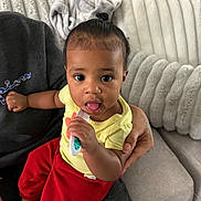 Ariyah joined the competition — help win amazing prizes! baby, child, person, toothbrush, couch, blanket, hand, red_pants, yellow_shirt, indoor, curly_hair, top_knot, plush_cushion, seated, cute, infant, looking_at_camera, comfort, home, caregiver