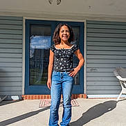Zola is registered to the contest to win money with this photo: girl, child, smile, porch, door, jeans, casual_clothing, sunlight, shadow, blue, outdoor, standing, young, person, sneakers, brick, concrete, wicker_chair, hand_on_hip, happy