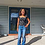 girl, child, smile, porch, door, jeans, casual_clothing, sunlight, shadow, blue, outdoor, standing, young, person, sneakers, brick, concrete, wicker_chair, hand_on_hip, happy