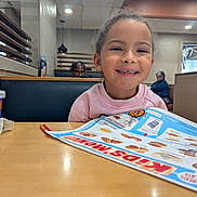 Naima is registered to the contest to win money with this photo: child, girl, smiling, restaurant, menu, table, cup, booth, indoor, happy, clothing, person, food, drink, dining, seat, menu_board, casual, family, portrait