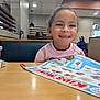 child, girl, smiling, restaurant, menu, table, cup, booth, indoor, happy, clothing, person, food, drink, dining, seat, menu_board, casual, family, portrait