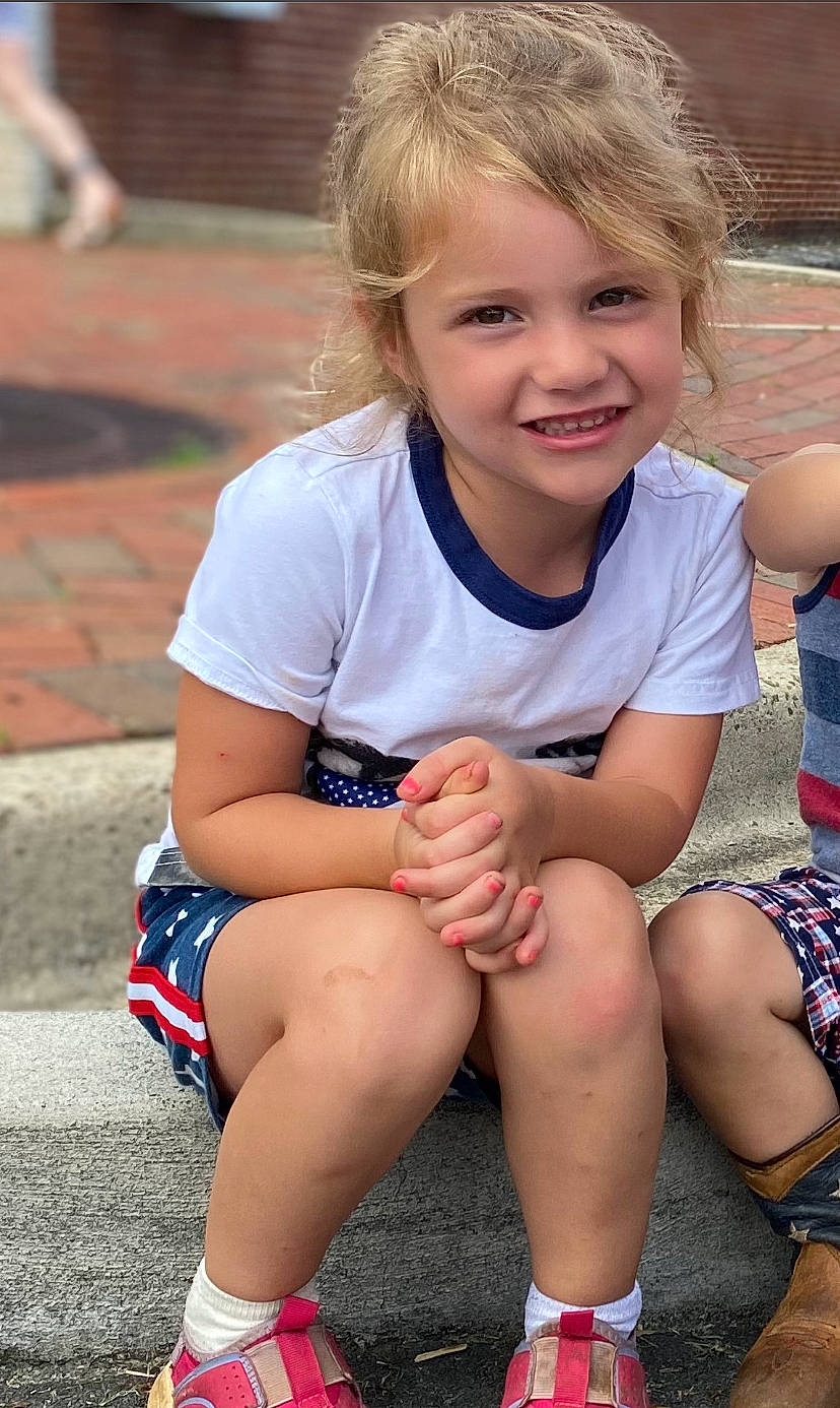 Ava is registered to the contest to win money with this photo: blond, child, electric_blue, face, facial_expression, fun, grass, happy, head, human_leg, joy, knee, leisure, person, shorts, shoulder, sitting, skin, smile, t_shirt