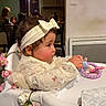 baby, bow, child, dining_room, floral_decor, hand, headband, highchair, indoor, kitchen, lace_dress, people_in_background, profile_view, rosy_cheeks, sitting, table, tablecloth, teether, toddler, toy