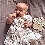 baby, infant, smiling, white_dress, patterned_clothing, lying_down, hand_in_mouth, cute, milestone_sign, 3_months, wooden_sign, flower_shape, bed_sheet, soft_fabric, happy, child, portrait, indoors, young_child, person