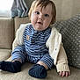 baby, child, infant, blue_eyes, striped_pajamas, socks, couch, indoor, curly_hair, cozy_clothing, cute, seated, portrait, soft_texture, home, furniture, curious_expression, warm_light, casual, smiling
