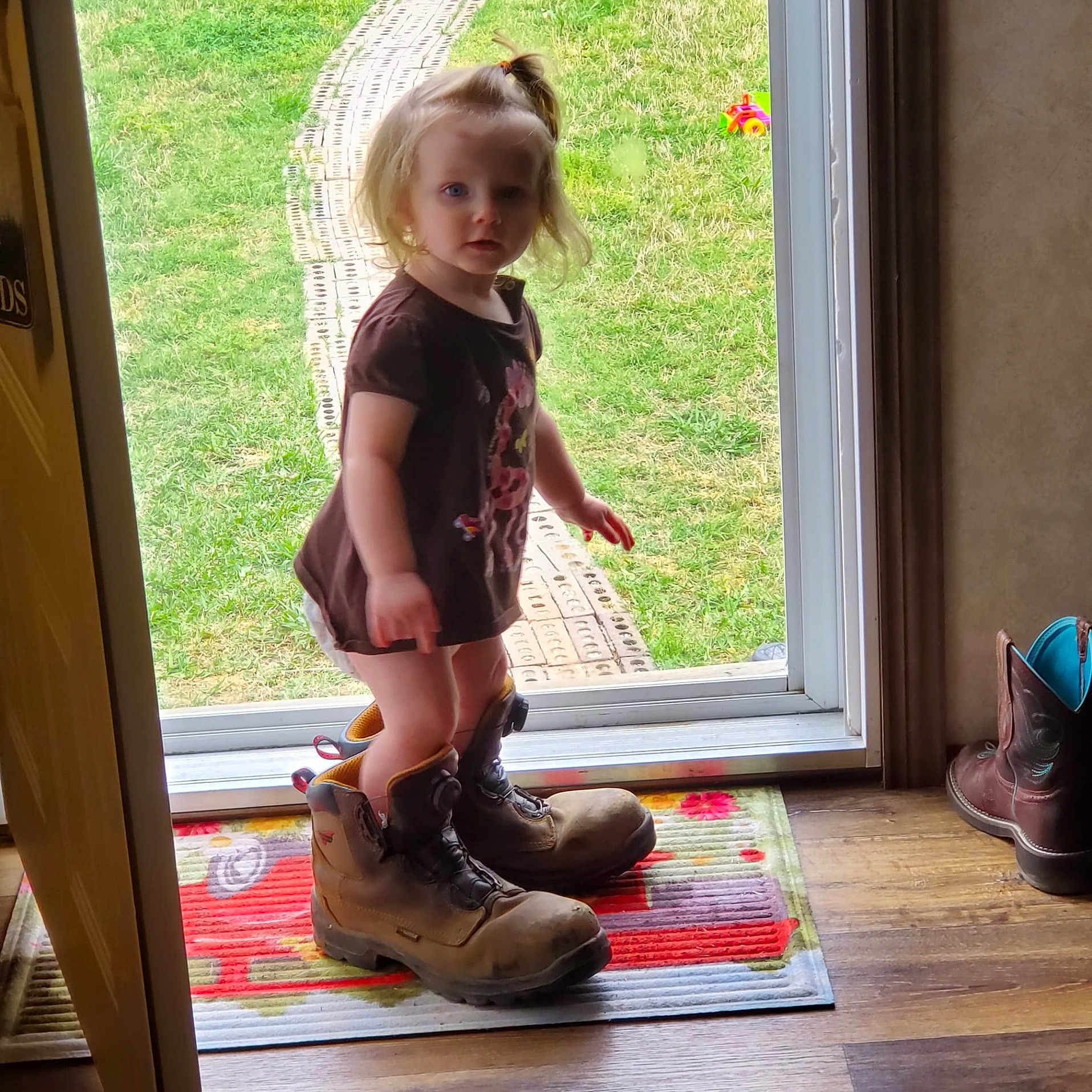 Lilly joined the competition — help win amazing prizes! child, toddler, girl, blonde_hair, ponytail, face, standing, outdoor, doorway, backyard, grass, pathway, boots, oversized_shoes, clothing, dress, toy, doormat, wooden_floor, portrait