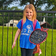 Lilly joined the competition — help win amazing prizes! child, girl, smiling, portrait, outdoor, school, backpack, chalkboard, sign, handwritten_text, blue_shirt, blonde_hair, standing, railing, garden, grass, trees, school_celebration, plush_toy, daylight