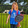 child, girl, smiling, portrait, outdoor, school, backpack, chalkboard, sign, handwritten_text, blue_shirt, blonde_hair, standing, railing, garden, grass, trees, school_celebration, plush_toy, daylight